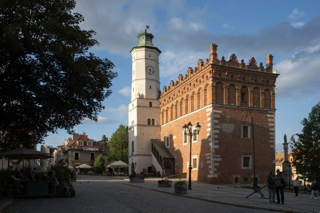 Renesansowy ratusz z białą wieżą zegarową i ozdobnym ceglanym budynkiem położony na głównym rynku, otoczony brukowaną ulicą oraz kilkoma grupami ludzi i kawiarnianymi ogródkami w cieniu drzew.