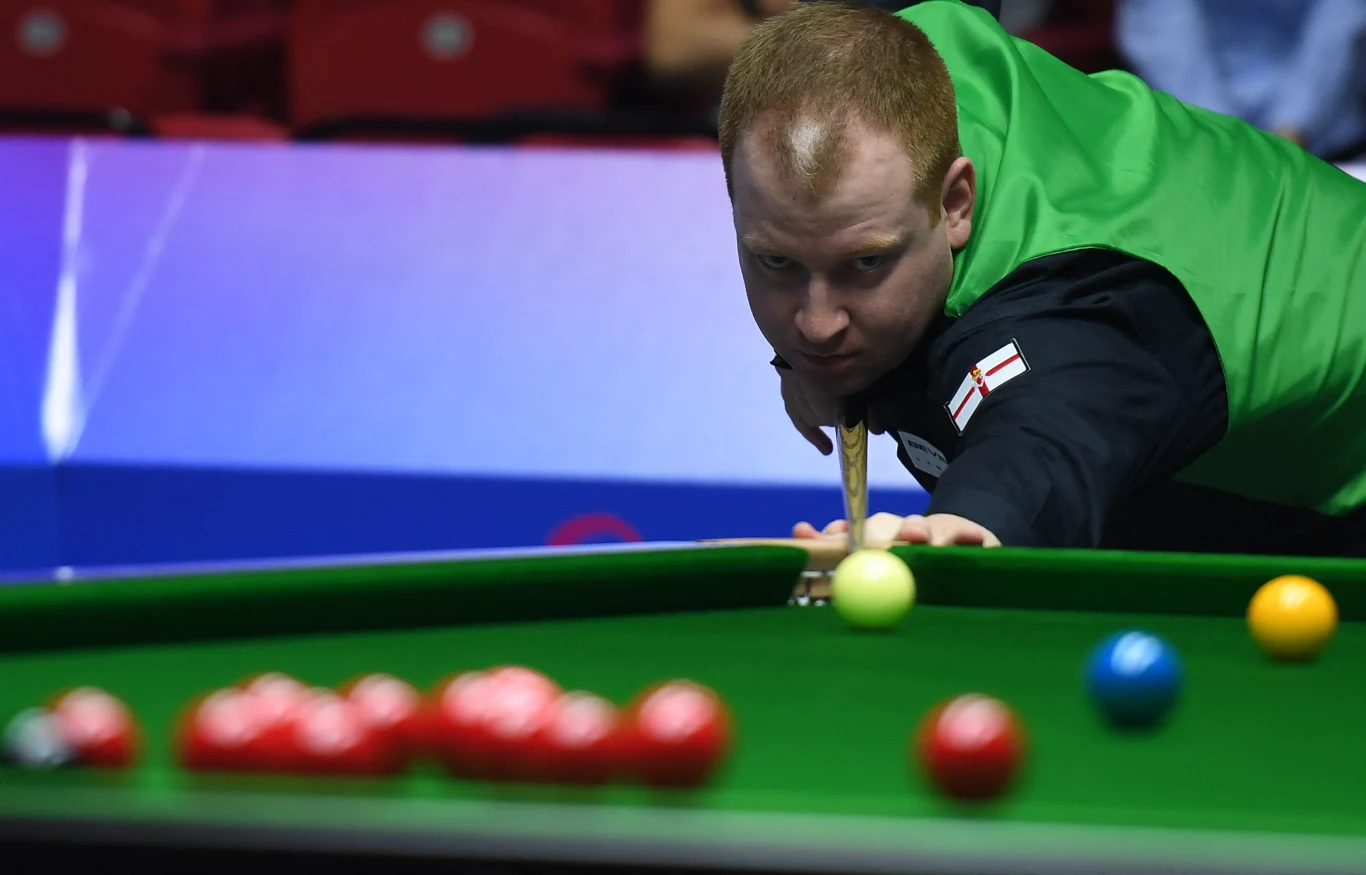Jordan Brown Zawodnik grający w snookera koncentruje się przy stole, przygotowując uderzenie kijem bilardowym. Stół pokryty zielonym suknem, na nim widoczne są bile różnego koloru, zawodnik ubrany jest w kamizelkę i koszulę, na rękawie znajduje się flaga Anglii.