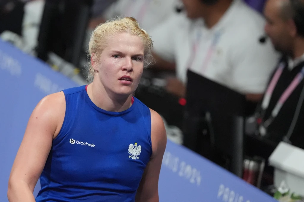 Elżbieta Wójcik Kobieta w niebieskim stroju sportowym z godłem Polski na piersi, stojąca na tle osób obsługujących wydarzenie sportowe; skupiona wyraz twarzy i krótkie blond włosy.