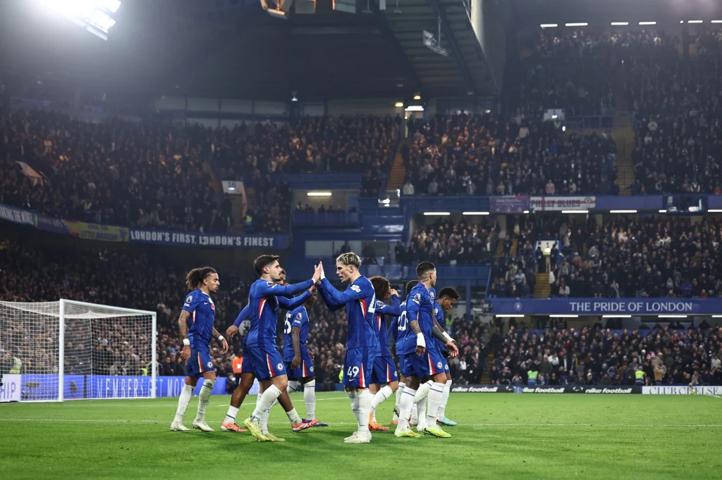 Piłkarze Chelsea Londyn w sezonie 2025/2026 Grupa piłkarzy w niebieskich strojach świętuje zdobycie bramki na murawie stadionu, w tle wypełnione trybuny z kibicami, widoczne napisy związane z klubem.