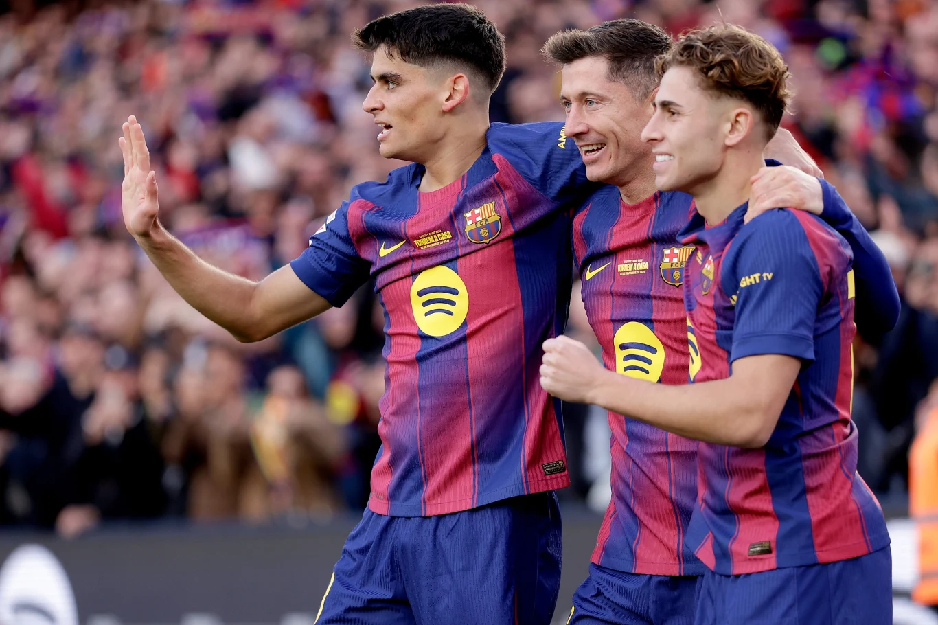 Gerard Martin, Robert Lewandowski i Fermin Lopez w barwach FC Barcelona Trzech zawodników w strojach FC Barcelony świętuje wspólnie na boisku, obejmując się ramionami, w tle widoczni kibice na trybunach.