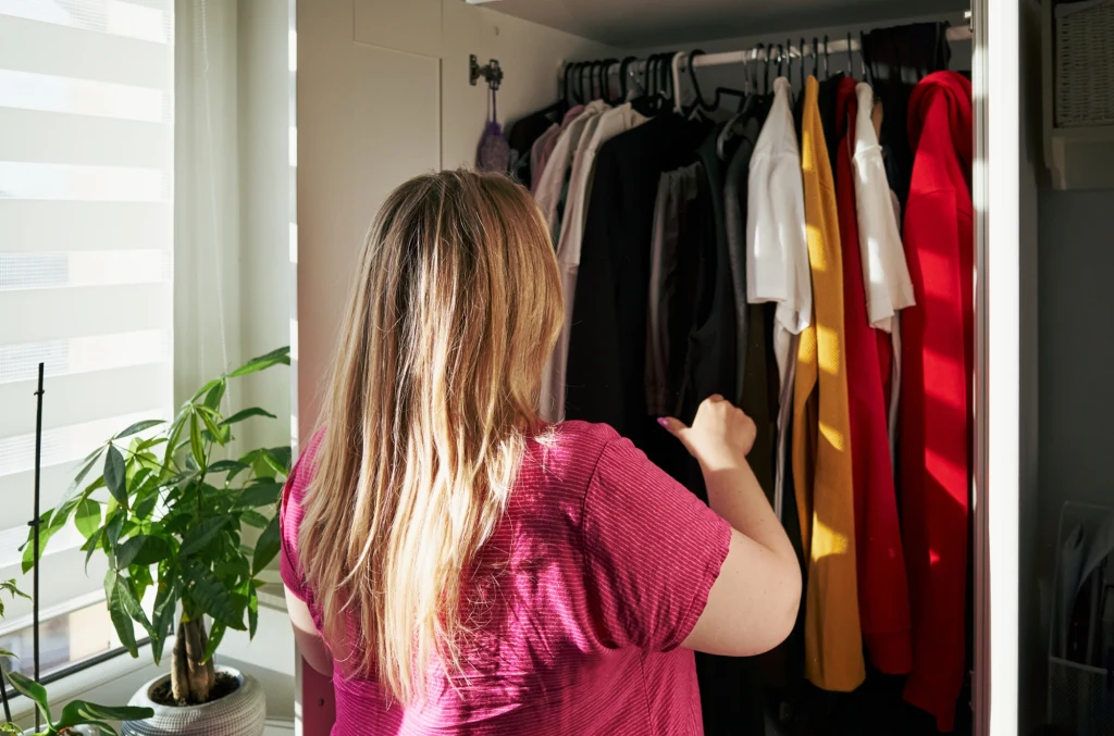 Otwierasz szafę i brzydko w niej pachnie? Spokojnie, można szybko pozbyć się problemu Kobieta z długimi blond włosami, ubrana w różową bluzkę, stoi tyłem i wybiera ubrania z szafy pełnej kolorowych ubrań. Obok szafy znajduje się roślina doniczkowa ustawiona przy oknie, przez które wpada dzienne światło.