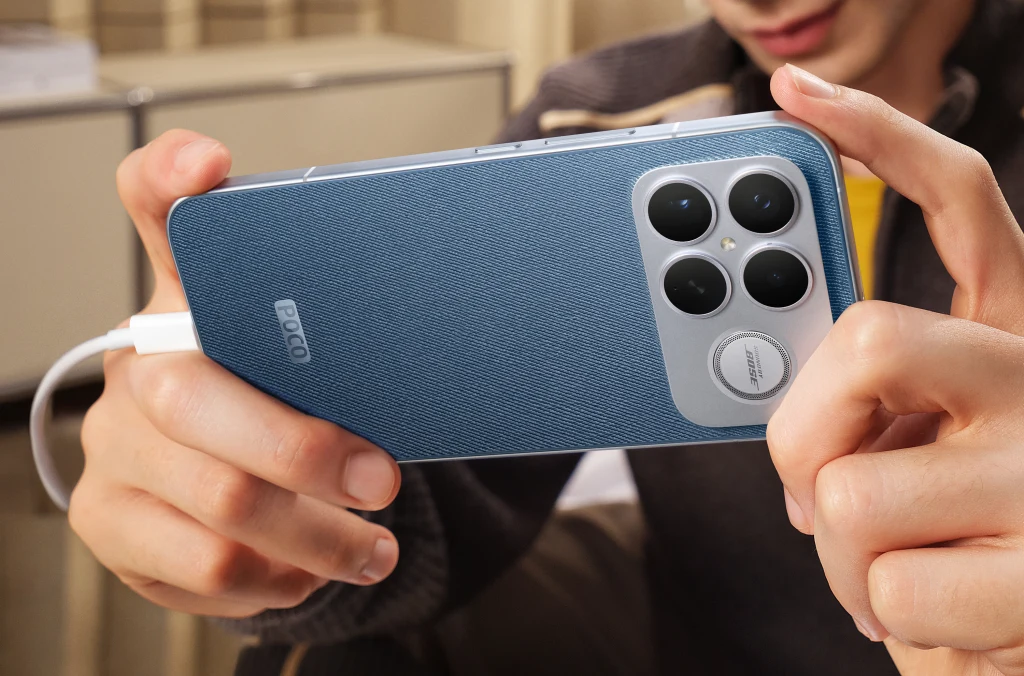 Młoda osoba trzyma niebieskiego smartfona marki POCO z dużym modułem aparatów i podłączonym przewodem do ładowania, koncentrując się na ekranie telefonu.