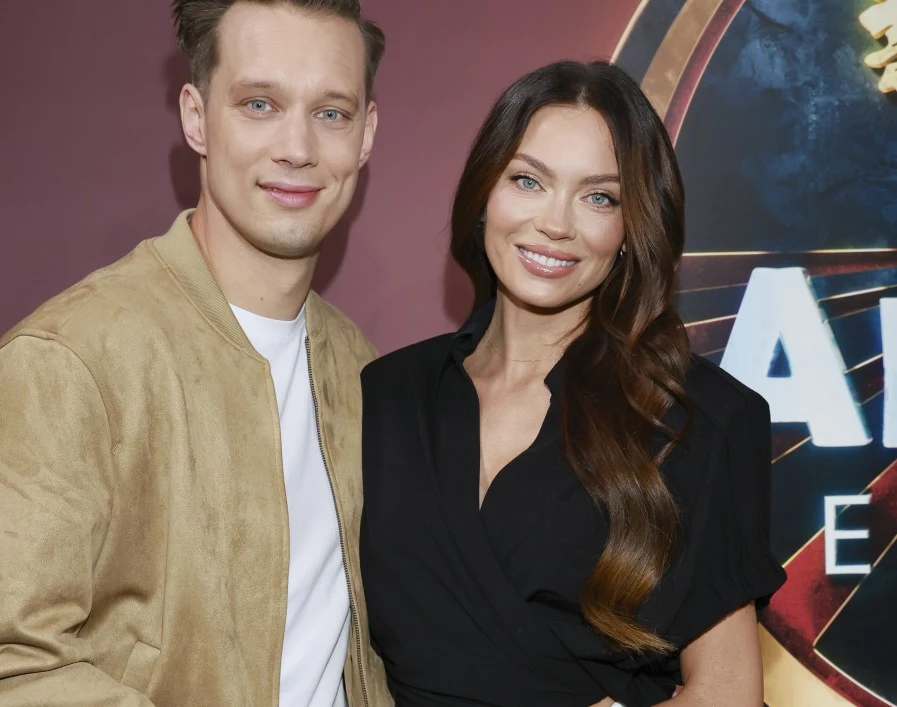 Uśmiechnięta kobieta z długimi, falowanymi włosami ubrana na czarno stoi obok mężczyzny w jasnej kurtce i białej koszulce, oboje pozują razem na tle dekoracyjnej ściany z motywem graficznym.