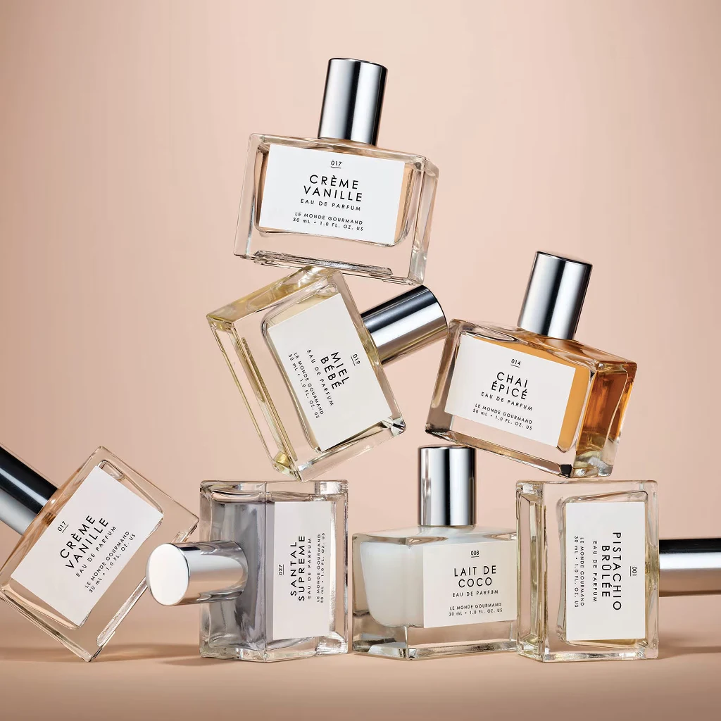 Zbiór siedmiu flakonów perfum o różnych nazwach i zapachach ułożonych w artystyczną kompozycję na beżowym tle. Każdy flakon ma prostokątny kształt, srebrną nakrętkę i białą etykietę z nazwą nut zapachowych.