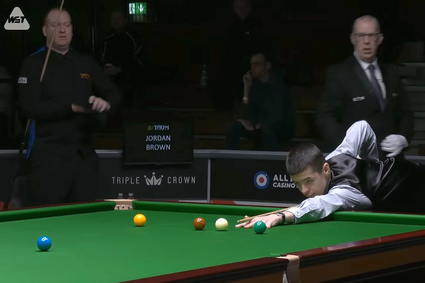 Michał Szubarczyk przy stole. Z lewej Jordan Brown Zawodnik snookera wykonuje precyzyjne uderzenie na stole, wokół znajdują się kolorowe bile oraz dwóch sędziów obserwujących przebieg gry, w tle plansza z nazwiskami uczestników meczu.