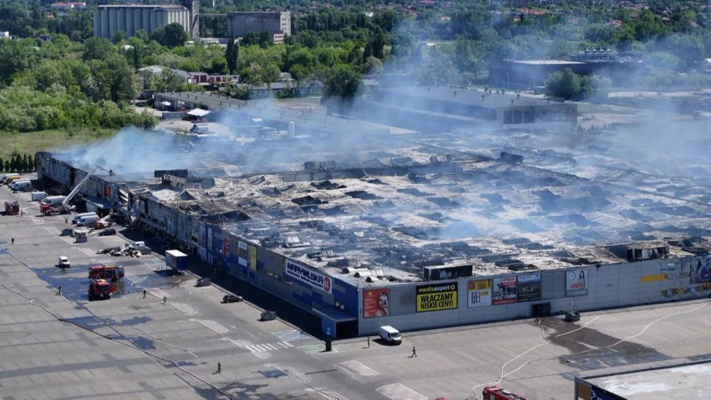 Pożar centrum handlowego "Marywilska 44" 12.05.2024 Spalony dach rozległego centrum handlowego, unoszący się dym i wozy strażackie pracujące wokół budynku; teren otoczony dużym parkingiem, w tle zieleń i zabudowania miejskie.