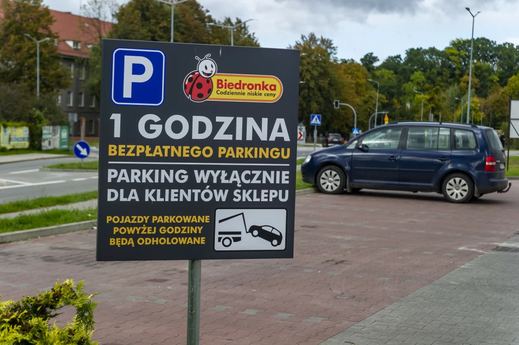 Tablica informacyjna sklepu Biedronka z informacją o godzinie bezpłatnego parkowania wyłącznie dla klientów oraz ostrzeżeniem o odholowywaniu pojazdów po przekroczeniu limitu czasu. W tle widoczny zaparkowany samochód na parkingu oraz fragment ulicy i ...