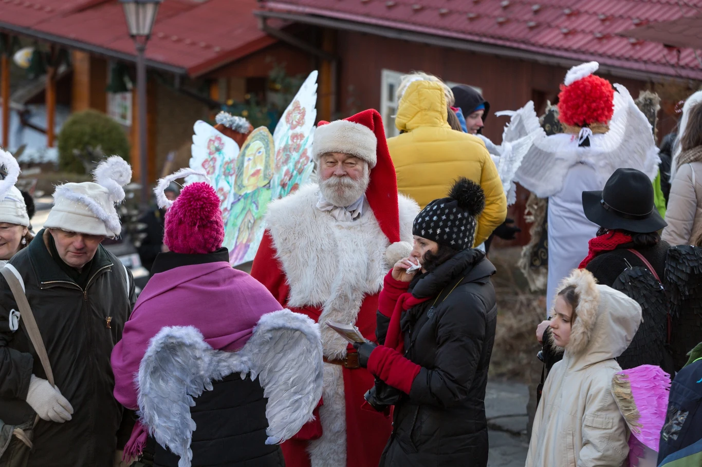 Grupa ludzi ubranych w kostiumy świąteczne, wśród których dominuje postać mężczyzny przebrana za Świętego Mikołaja. W tle osoby w przebraniach aniołów z białymi skrzydłami, kurtkami i czapkami chroniącymi przed zimnem, rozmowy i świąteczna atmosfera na...