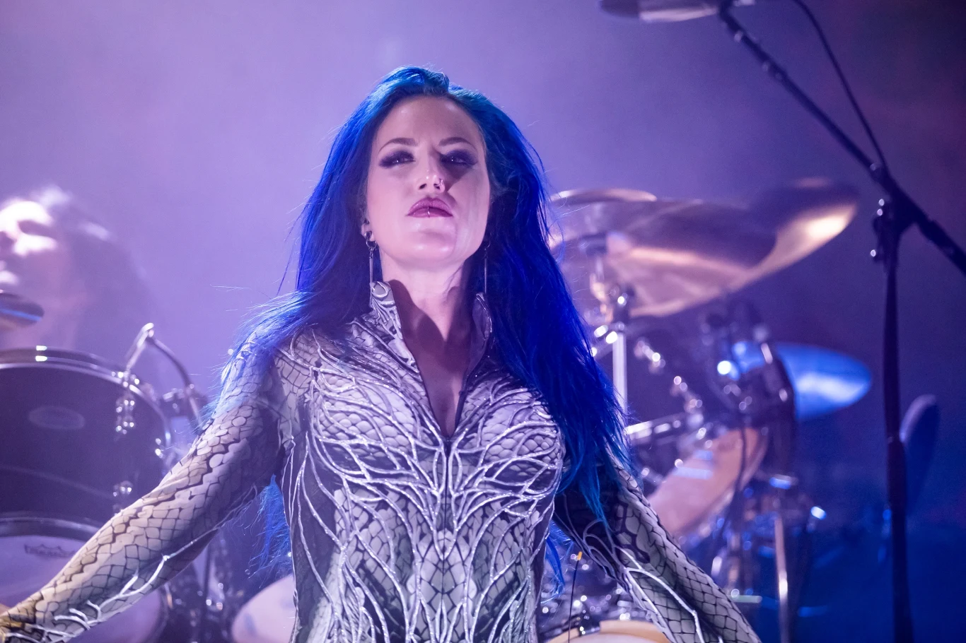 Alissa White-Gluz po 12 latach rozstała się z grupą Arch Enemy Wokalistka o niebieskich włosach stoi na scenie w świetle reflektorów, w tle widoczne są bębny i elementy perkusji. Jej wyraz twarzy jest dumny i pewny siebie, a stroje sceniczne przyciągają uwagę wzorami nawiązującymi do motywów organicznych.