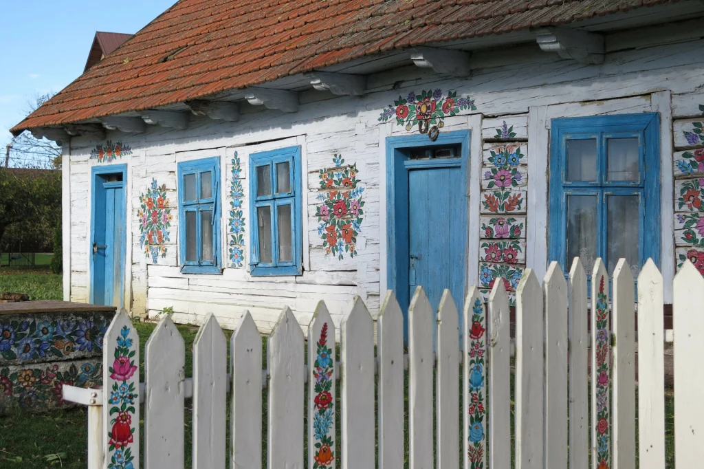 Zalipie to jedyna taka wieś w Polsce Stara drewniana chałupa z niebieskimi oknami i drzwiami, ozdobiona kolorowymi kwiatowymi wzorami na ścianach i białym, pomalowanym płocie; w tle widoczna część ogrodu.