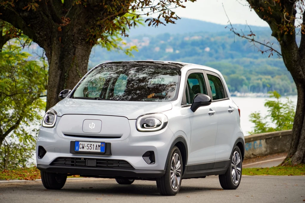 Leapmotor T03 z zewnątrz kojarzy się ze Smartem EQ ForFour Biały, kompaktowy samochód elektryczny zaparkowany przy asfaltowej drodze, w tle jezioro, drzewa oraz wzgórza porośnięte zielenią. Auto posiada włoską tablicę rejestracyjną, a za nim widoczna jest betonowa bariera przy zbiorniku wodnym.