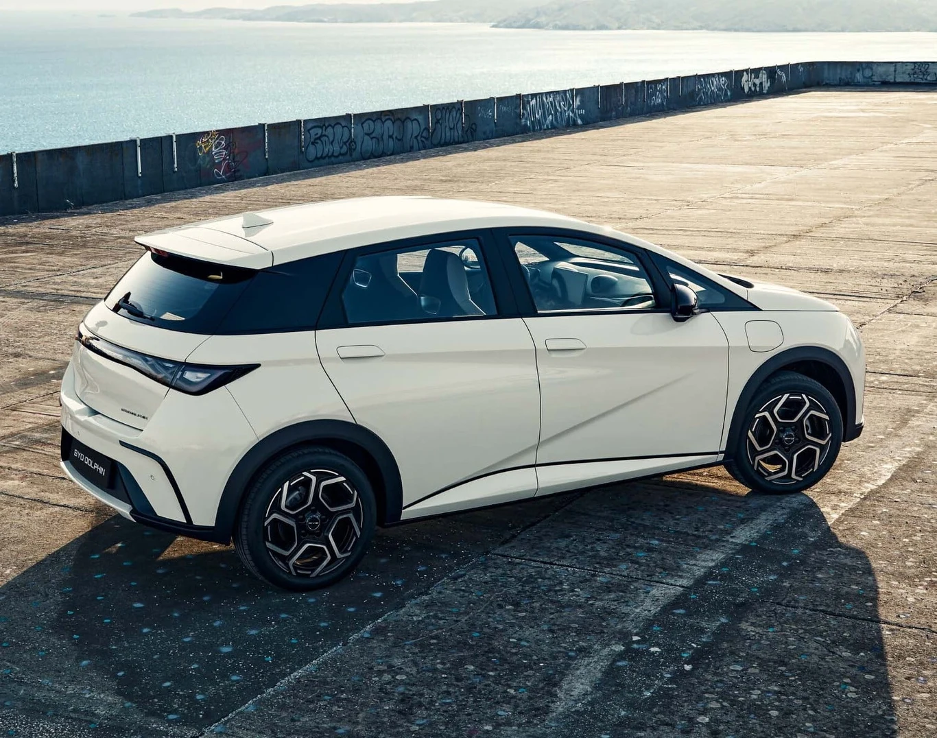 Nowoczesny, biały samochód osobowy zaparkowany na betonowym placu z widokiem na wodę i muralami na murze w tle.