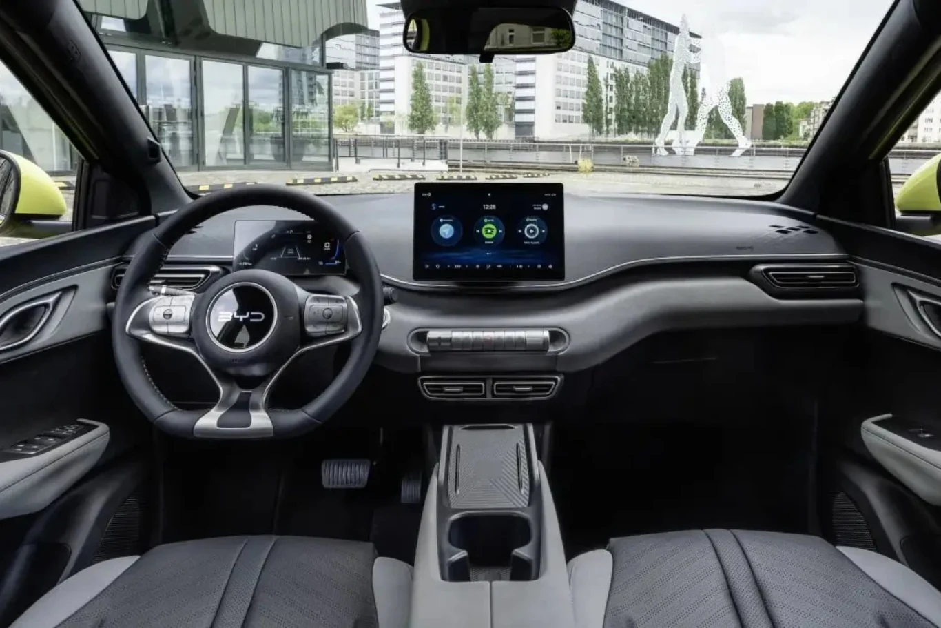 Nowoczesna kabina samochodu z dużym, centralnym wyświetlaczem dotykowym, cyfrowym zegarami za kierownicą i minimalistycznym wykończeniem, przez przednią szybę widać elementy architektury miejskiej oraz szklaną rzeźbę sylwetki człowieka.