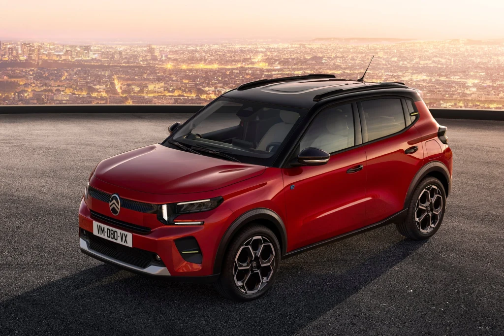 Citroen e-C3 to elektryczny wariant doskonale znanego modelu C3 Czerwony samochód Citroën C3 Aircross zaparkowany na tle panoramy miasta o zachodzie słońca, nowoczesny design, kontrastujący czarny dach, widoczne charakterystyczne felgi oraz eleganckie reflektory.