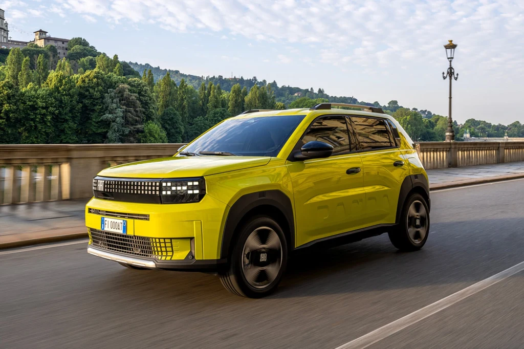 Fiat Grande Panda zastąpił modele Panda i Punto Jaskrawożółty samochód SUV jadący po szerokiej ulicy mostu, w tle rozległy zielony park oraz stylowe, historyczne budynki. Pojazd wyróżnia się nowoczesnym designem i dynamiczną sylwetką, a drogę otacza klasyczna latarnia miejska.