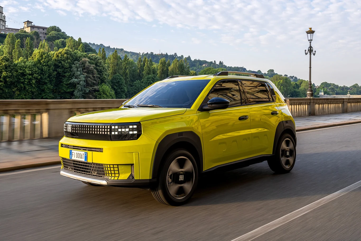 Jaskrawożółty samochód SUV jadący po szerokiej ulicy mostu, w tle rozległy zielony park oraz stylowe, historyczne budynki. Pojazd wyróżnia się nowoczesnym designem i dynamiczną sylwetką, a drogę otacza klasyczna latarnia miejska.