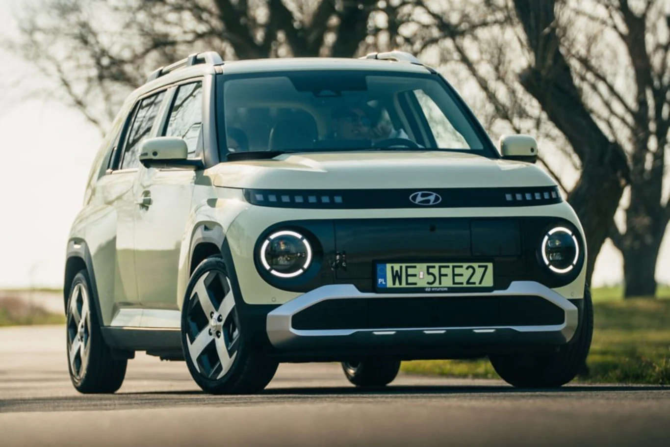 Nowoczesny samochód Hyundai o jasnej karoserii zaparkowany na asfalcie, widoczny z przodu na tle natury i drzew, wyraźne okrągłe reflektory oraz miejska rejestracja.