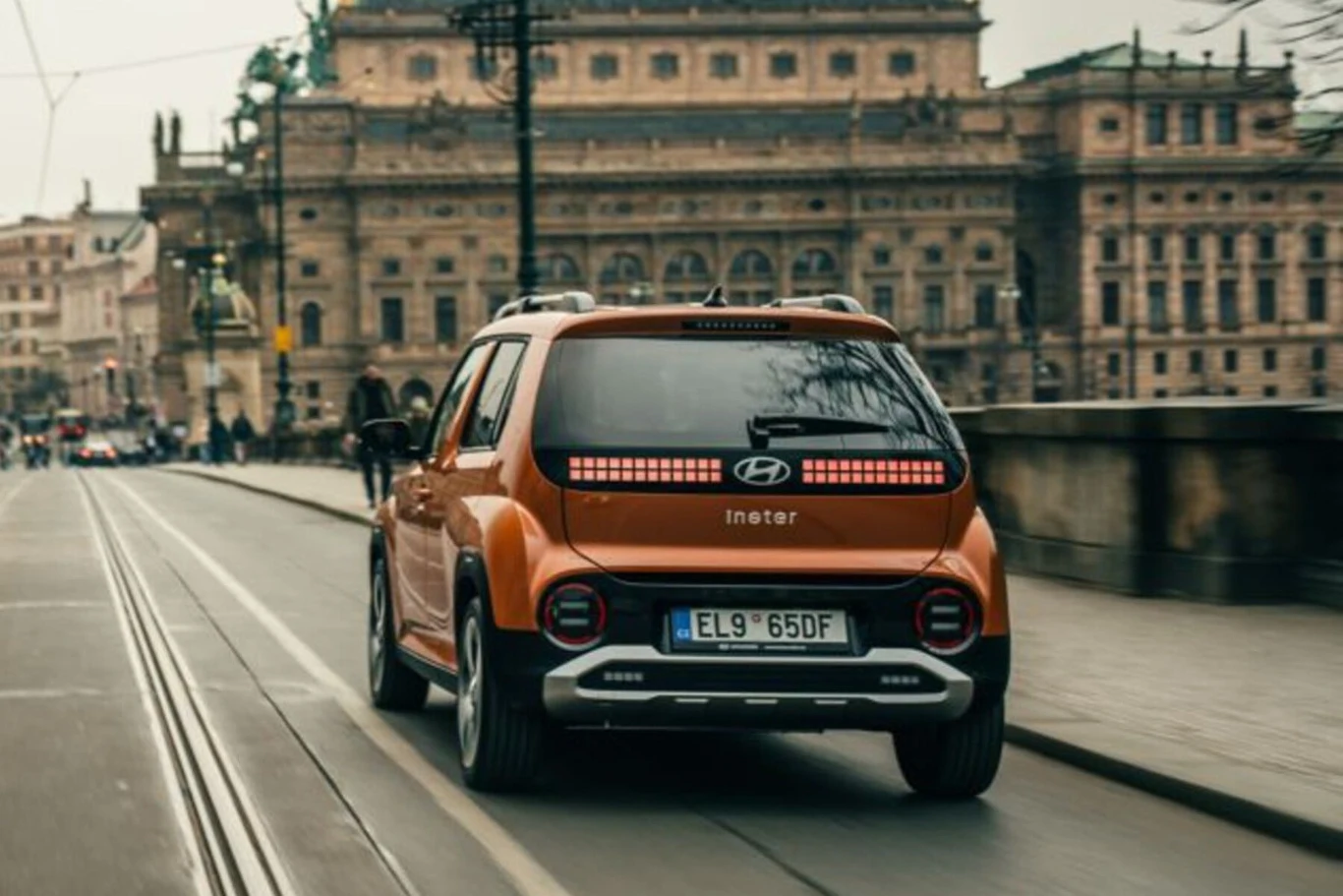 Pomarańczowy samochód marki Hyundai Inster jedzie ulicą w mieście, po lewej widoczne są tory tramwajowe, w tle znajduje się historyczny budynek o bogatej architekturze.
