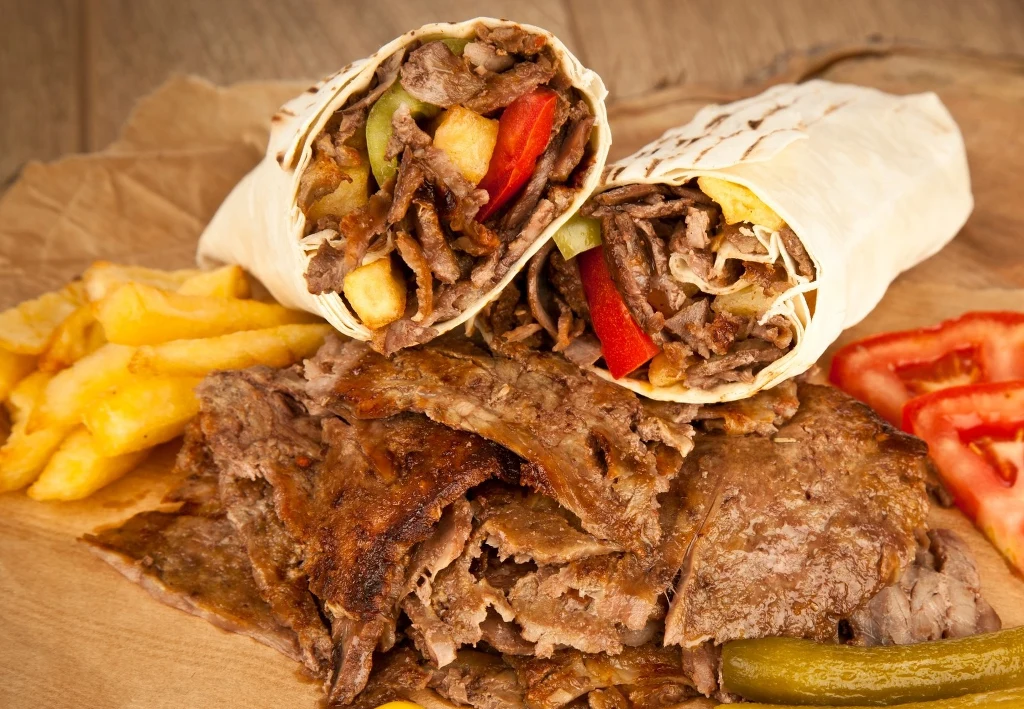 Placki tortilli z kawałkami mięsa kebab, frytkami, ogórkiem konserwowym oraz plasterkami świeżego pomidora na papierze.