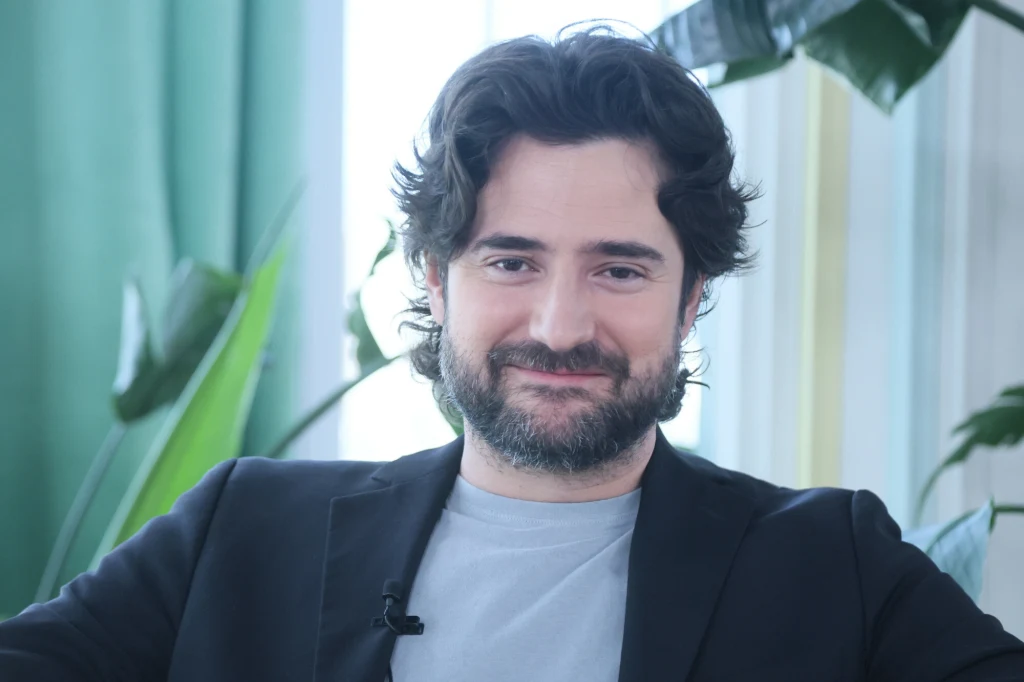 Uśmiechnięty mężczyzna z ciemnymi włosami i brodą, ubrany w ciemną marynarkę i jasny t-shirt, siedzi na tle jasnego wnętrza z zielonymi roślinami.