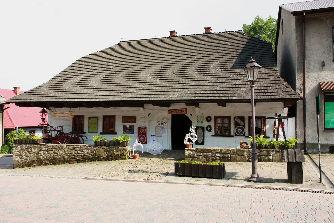 Drewniany dom z białymi ścianami i stromym dachem, ozdobiony kwiatami i tablicami informacyjnymi przy wejściu, otoczony kamiennym murkiem i brukowaną uliczką, obok znajduje się latarnia uliczna.