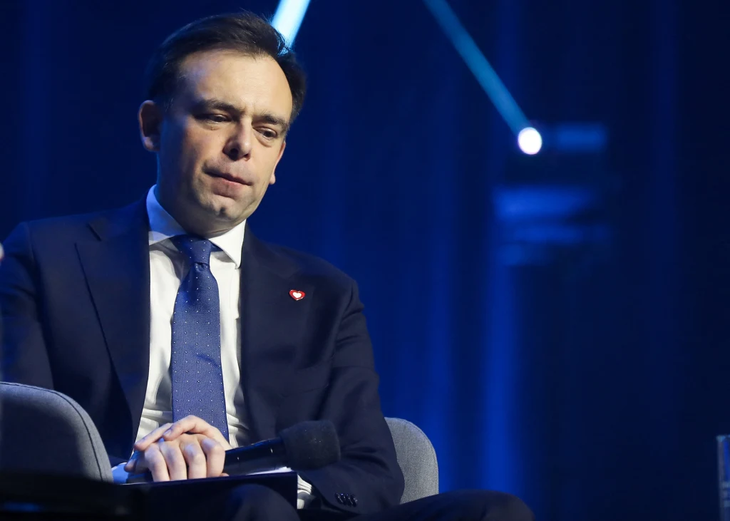 Mężczyzna w garniturze i krawacie siedzi na scenie, ze spuszczoną głową i zamyślonym wyrazem twarzy, trzyma mikrofon w dłoniach; w tle wyraźne niebieskie światło sceny.