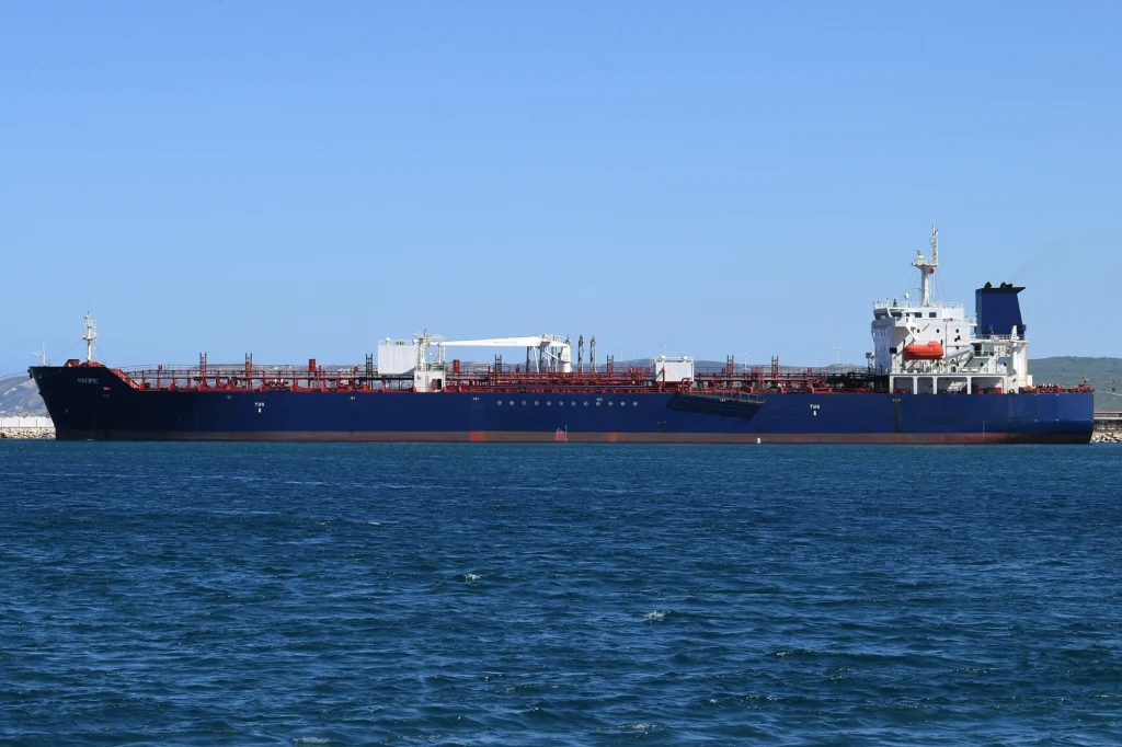 Duży tankowiec płynący po spokojnej wodzie w słoneczny dzień, niebo bez chmur, widoczna część lądu w tle.