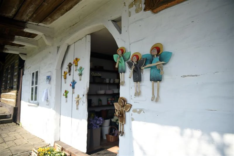 Biała, drewniana ściana chaty z zawieszonymi dekoracyjnymi aniołkami z materiału i ceramiki, otwarte drzwi prowadzą do wnętrza domu wypełnionego naczyniami, obok wejścia ustawione są kwiaty.