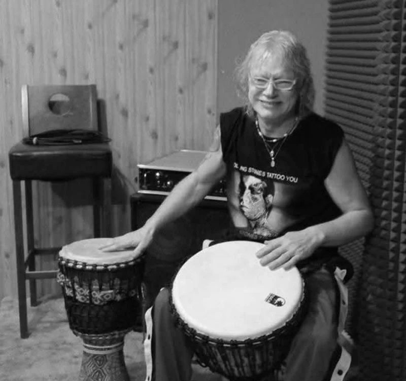 Janusz Staszek zmarł po ciężkiej chorobie Kobieta z uśmiechem grająca na dwóch bębnach djembe, siedząca w pomieszczeniu wyciszonym, obok stoi krzesło i sprzęt elektroniczny.