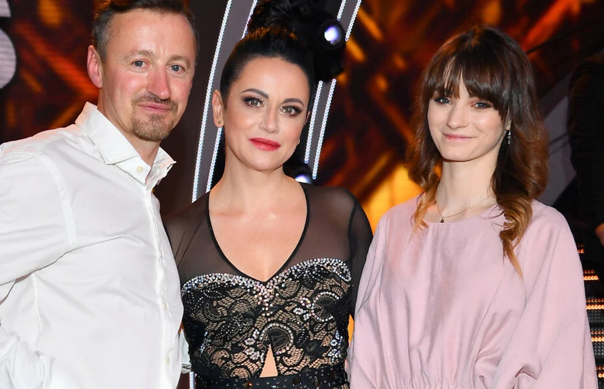 Adam, Izabela i Karolina Małyszowie Trzy osoby stoją obok siebie, środkowa kobieta ubrana w czarną, ozdobną suknię z siateczką, po jej lewej stronie mężczyzna w białej koszuli, po prawej kobieta w jasnoróżowej sukience, za nimi kolorowe, rozświetlone tło.