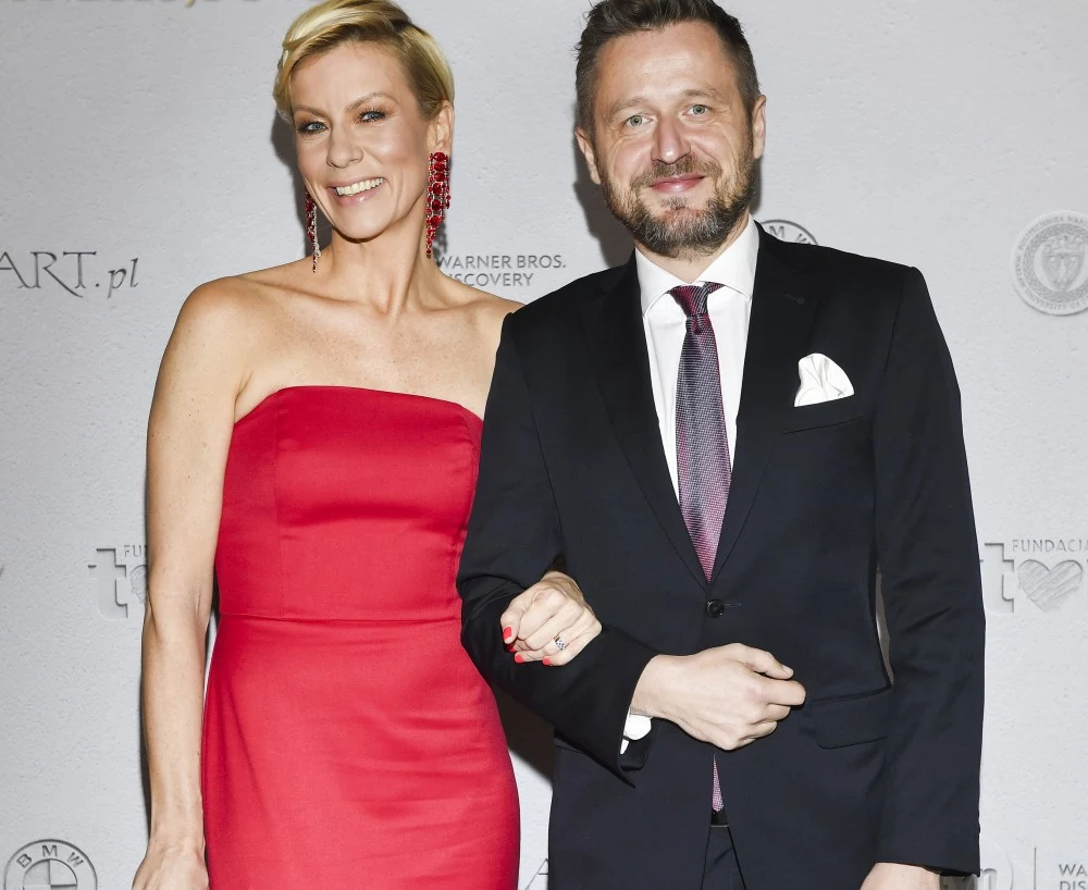 Elegancka kobieta w czerwonej sukni i mężczyzna w garniturze pozują razem na tle ściany z logotypami. Oboje uśmiechnięci, kobieta trzyma mężczyznę pod ramię. Oświetlenie profesjonalne, atmosfera formalna.