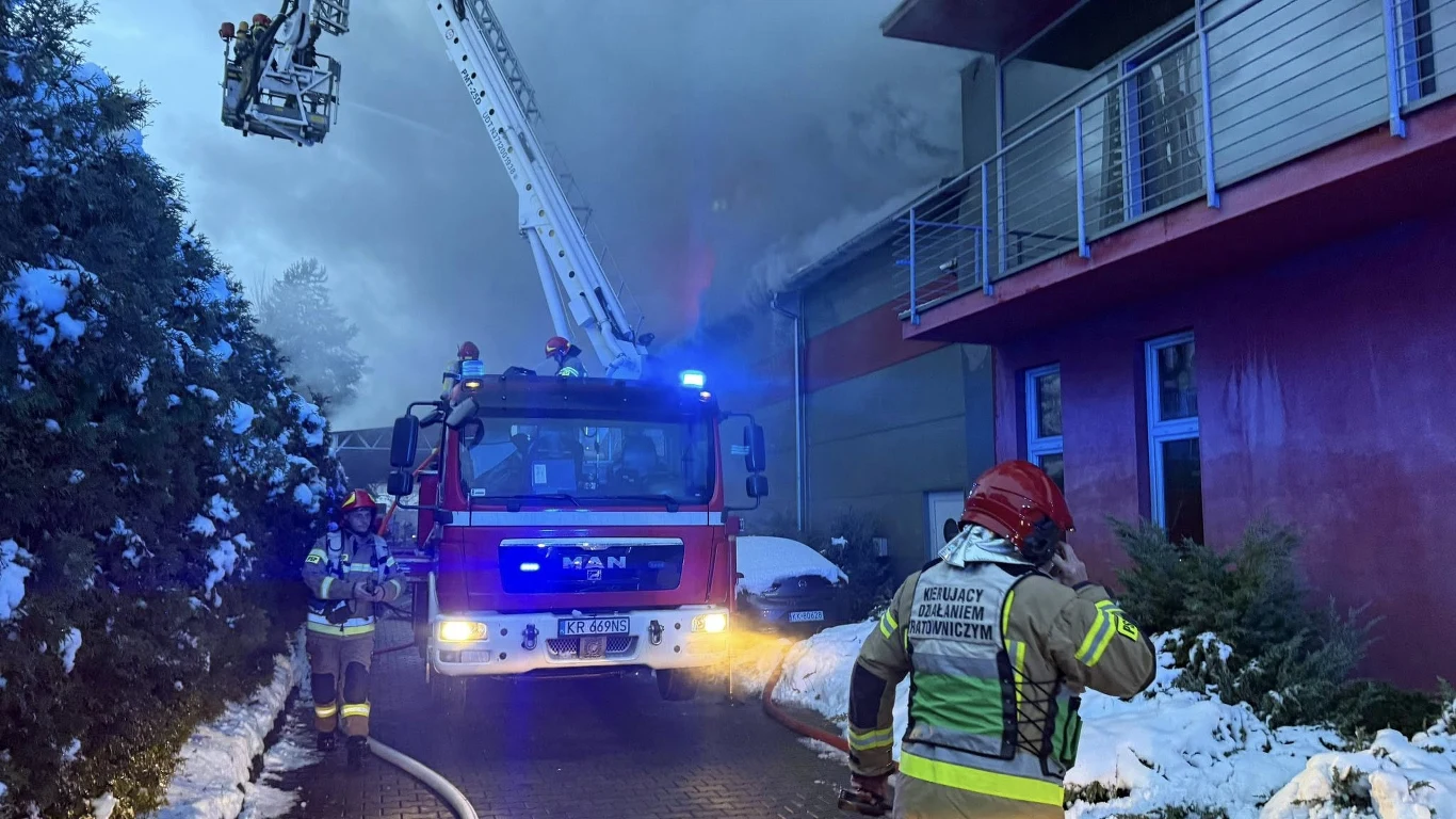 Pożar hali magazynowej w Skawinie pod Krakowem Strażacy w trakcie akcji gaśniczej przy budynku objętym pożarem, widoczny wóz strażacki z rozłożoną drabiną oraz gęsty dym wydobywający się z górnej części budynku, otoczenie pokryte śniegiem.