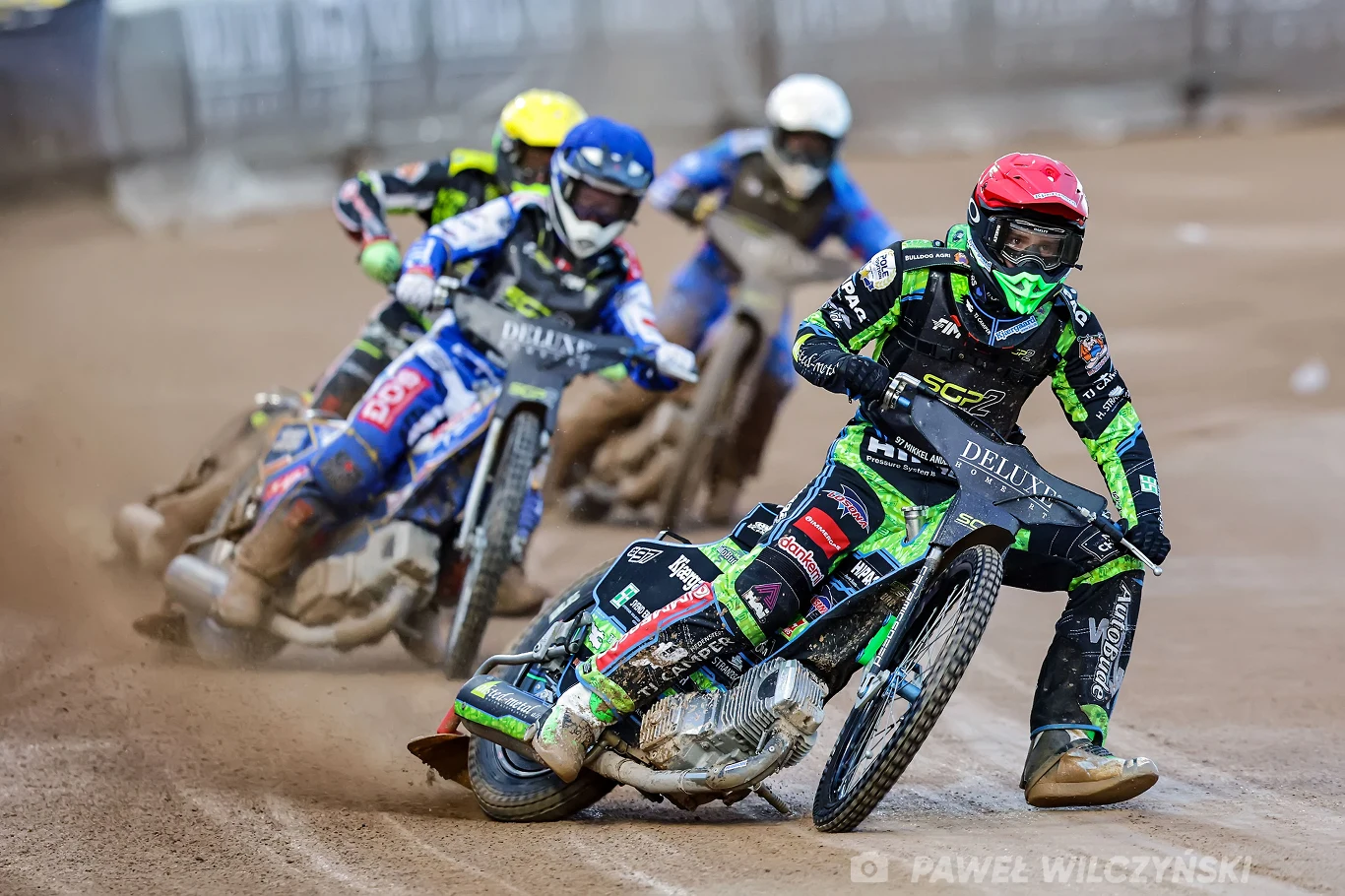 Mikkel Andersen na prowadzeniu Zawodnicy żużlowi w dynamicznym wyścigu na torze, widoczne motocykle ślizgające się po ziemi, unoszący się pył oraz sportowcy w kolorowych kaskach i kombinezonach.