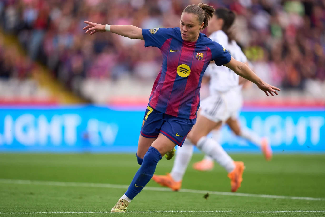 Ewa Pajor celebrująca bramkę Piłkarka FC Barcelona w niebiesko-czerwonym stroju wykonuje dynamiczny ruch kopiąc piłkę na boisku, w tle widoczni rywale w białych strojach oraz rozmyte trybuny z kibicami.
