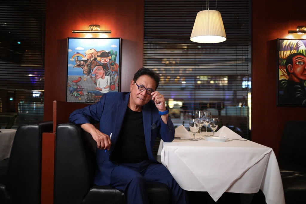 Mężczyzna w granatowym garniturze siedzi przy stole w eleganckiej restauracji, oparty wygodnie, w ręku trzyma okulary. Na stole przykrytym białym obrusem znajdują się kieliszki i nakrycia. W tle widoczne są kolorowe obrazy na ścianach oraz lampy sufitowe.