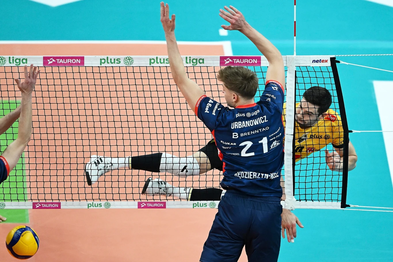Dwóch siatkarzy w dynamicznej akcji przy siatce, jeden z zawodników w żółtym stroju uderza piłkę, drugi w granatowym wykonuje blok. Widoczne są również fragmenty boiska i siatki.
