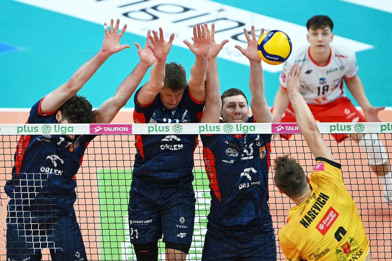 Trzech zawodników drużyny ubranej na granatowo wykonuje blok przy siatce przeciwko atakującemu graczowi w żółtej koszulce. Na drugim planie zawodnik w białej koszulce obserwuje akcję.