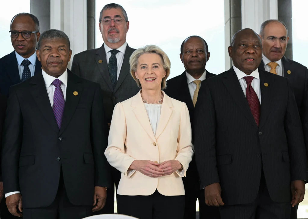 Grupa elegancko ubranych osób w garniturach i marynarce pozuje razem w jasnym pomieszczeniu, centralną postacią jest kobieta w kremowej marynarce, a pozostali to mężczyźni ubrani formalnie z przypiętymi odznakami.