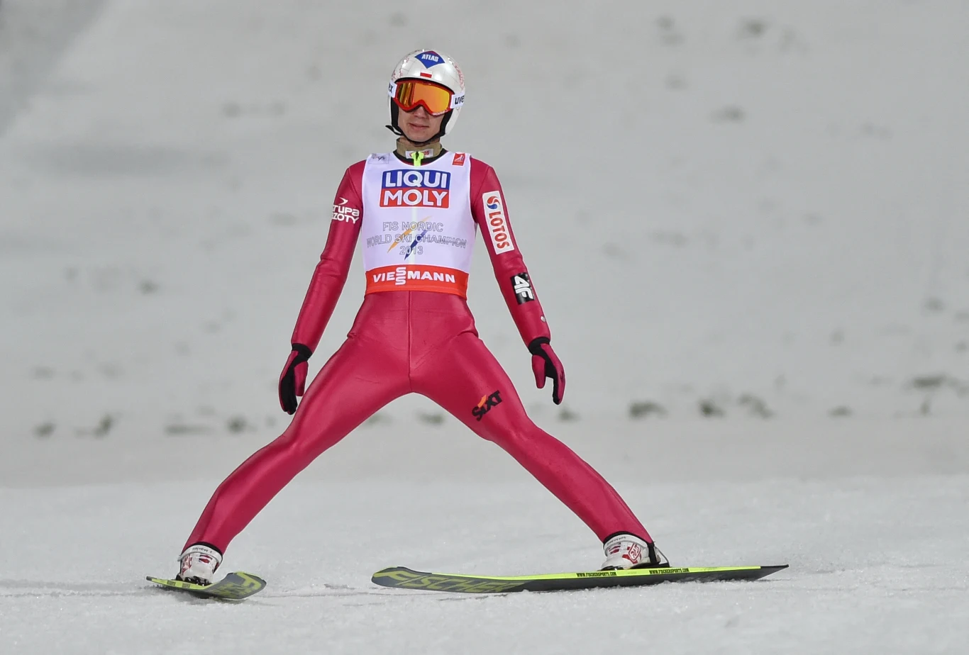 Kamil Stoch w czasie MŚ w Falun (2015 r.) Zawodnik w trakcie lądowania podczas skoku narciarskiego na śnieżnym zeskoku, ubrany w czerwony kombinezon startowy, kask i gogle ochronne, z wyciągniętymi szeroko nartami w charakterystycznej pozycji.