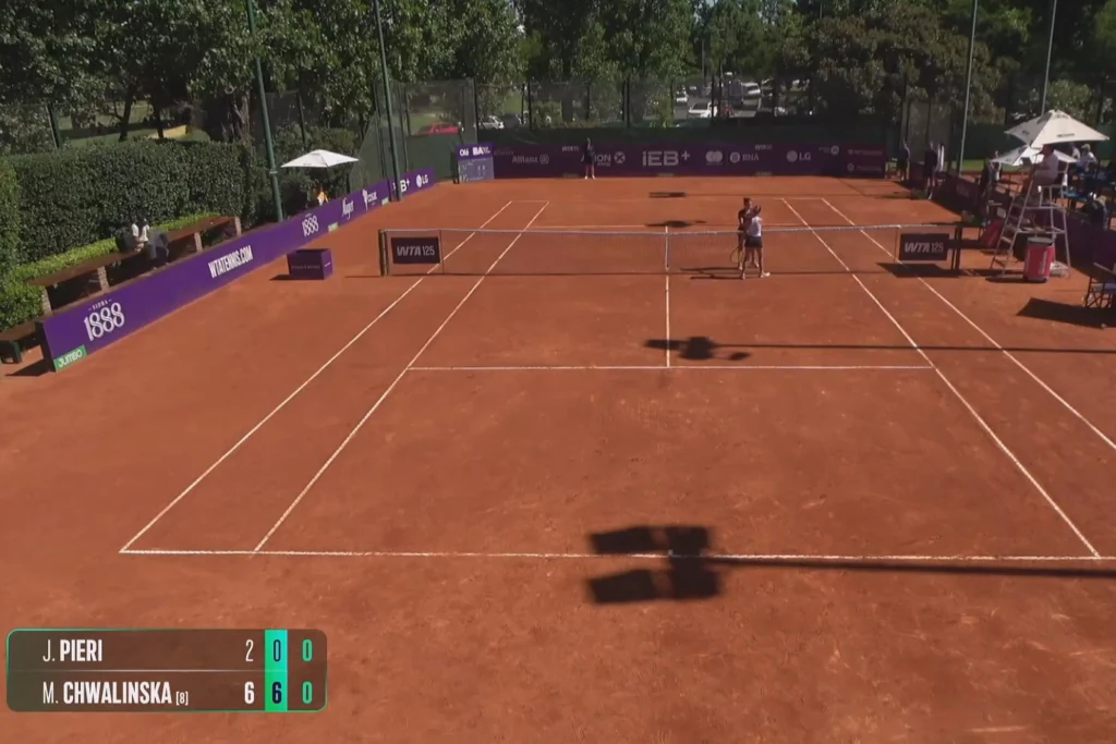 Kort tenisowy ziemny otoczony zielenią i trybunami, na środku dwie tenisistki przygotowujące się do gry, na dole widoczne wyniki z nazwiskami zawodniczek oraz przewaga jednej z nich, otoczenie turniejowe z bandami reklamowymi.