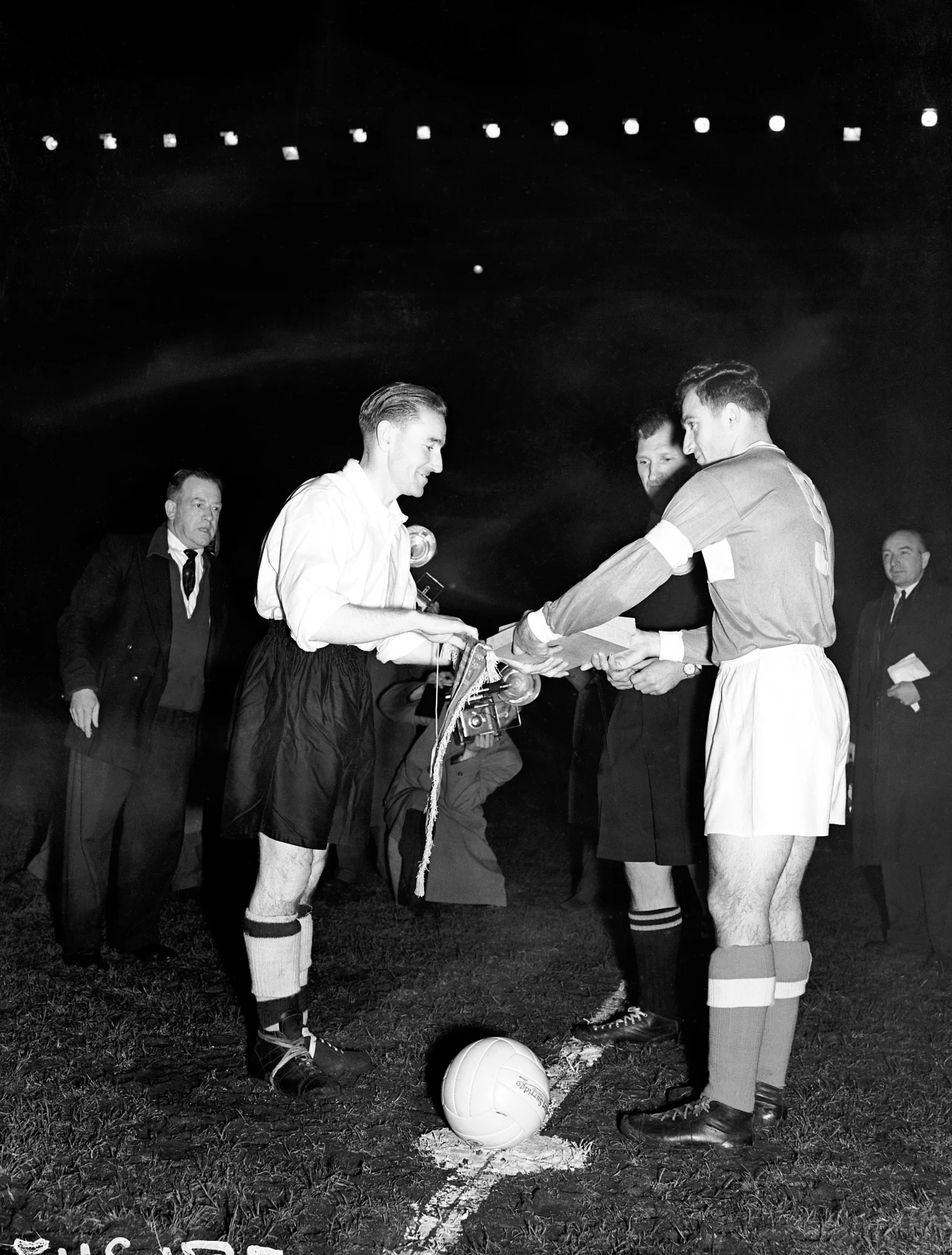 Dwóch piłkarzy w sportowych strojach stoi naprzeciwko siebie i wymienia proporczyki przed rozpoczęciem meczu. Sędzia obserwuje tę scenę z bliska, a w tle widoczne są sylwetki kilku innych mężczyzn oraz piłka na murawie. Światła stadionu podkreślają wie...