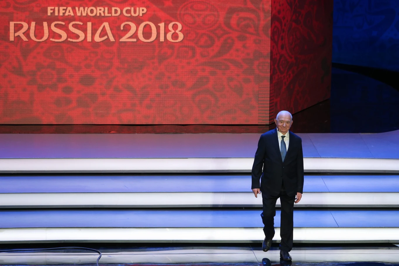 Mężczyzna w garniturze stoi na białych schodach sceny, w tle duży czerwony ekran z napisem 'FIFA World Cup Russia 2018'.