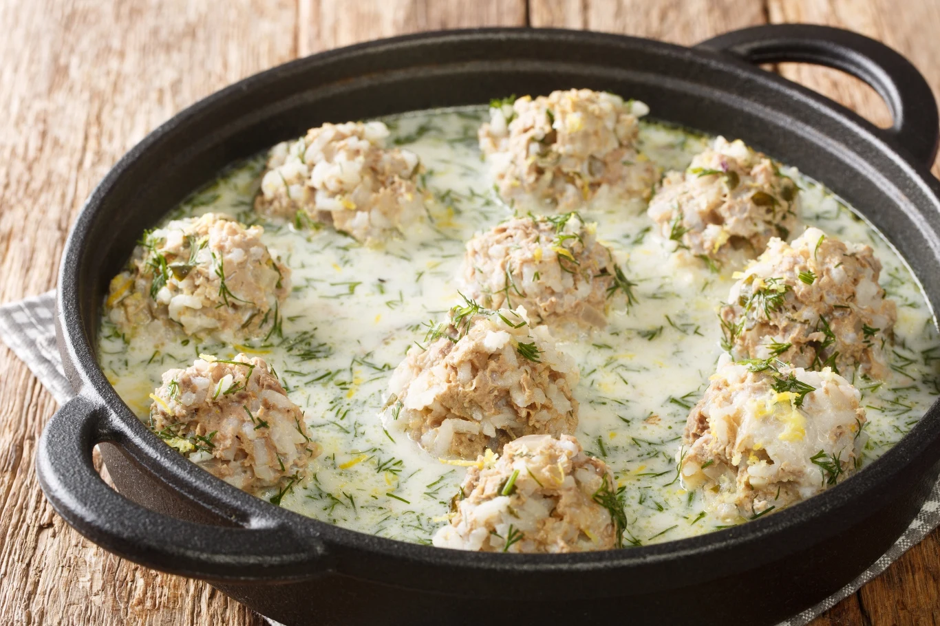 Okrągłe pulpety z mięsa i ryżu zanurzone w jasnym, kremowym sosie ziołowym, posypane świeżym koperkiem, podane w czarnym, żeliwnym naczyniu na drewnianym stole.