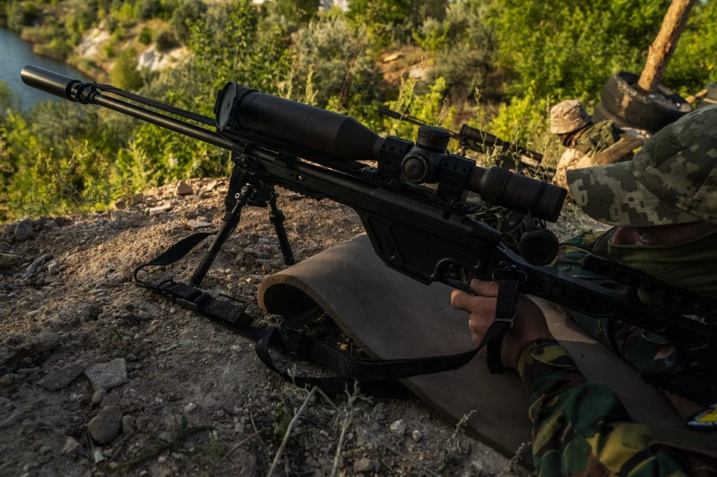 Snajper w mundurze maskującym i w kasku leży na ziemi przy krawędzi wzniesienia, celując przez lunetę karabinu z dużym obiektywem. W tle widoczna zielona roślinność i jezioro.