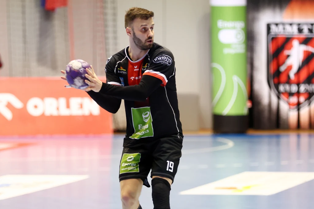 Michał Potoczny (MMTS Kwidzyn) Szczypiornista w czarnym stroju sportowym trzyma piłkę do piłki ręcznej, przygotowując się do podania lub rzutu podczas meczu na hali sportowej. W tle widoczne są reklamy oraz baner klubu sportowego.