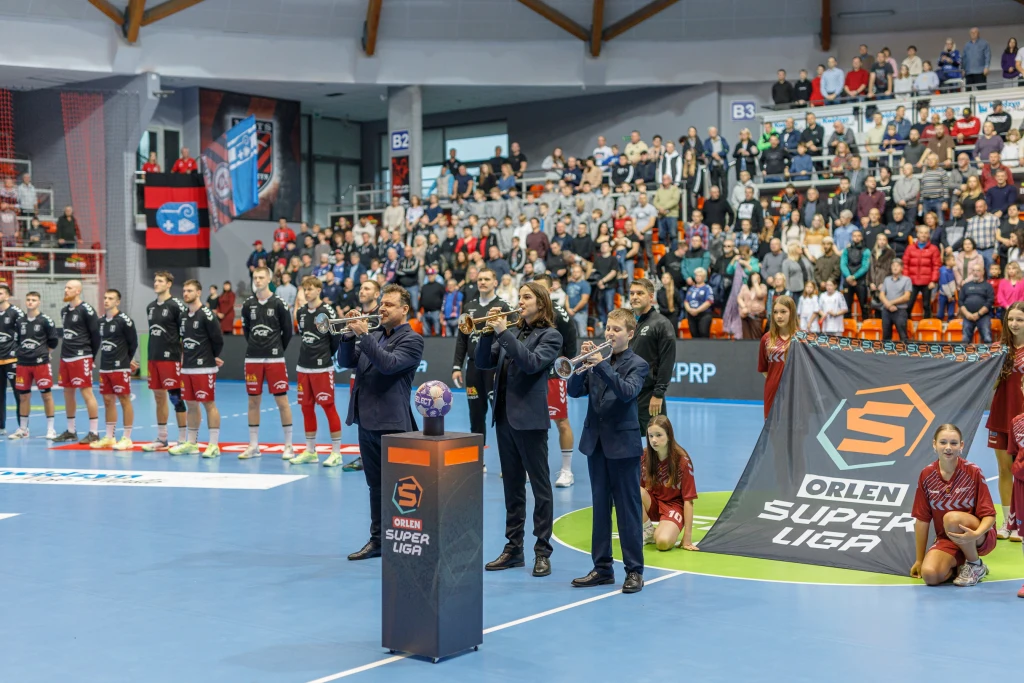 Grupa muzyków grających na trąbkach podczas ceremonii otwarcia meczu piłki ręcznej w hali sportowej, w tle ustawione drużyny sportowców w czarnych i czerwonych strojach, obok baner Orlen Superliga, na trybunach widzowie.