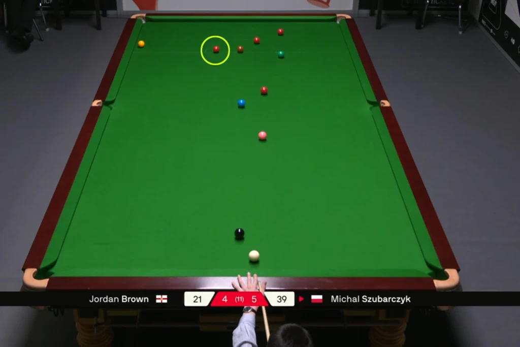 Stół do snookera podczas trwającej rozgrywki, widoczne bile w różnych kolorach, gracz przygotowujący się do wykonania uderzenia, na dole ekranu pasek wyników z nazwiskami zawodników i aktualnym stanem meczu.