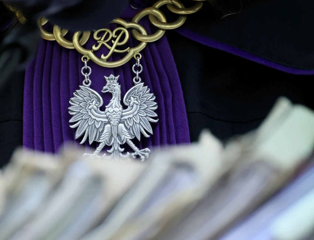 Nakaz alimentacyjny w planach rządu. Sądy zyskają nowe uprawnienia Orzeł biały w koronie na łańcuchu urzędowym zawieszonym na szyi osoby w todze sędziowskiej, na pierwszym planie rozmazane akta sądowe.
