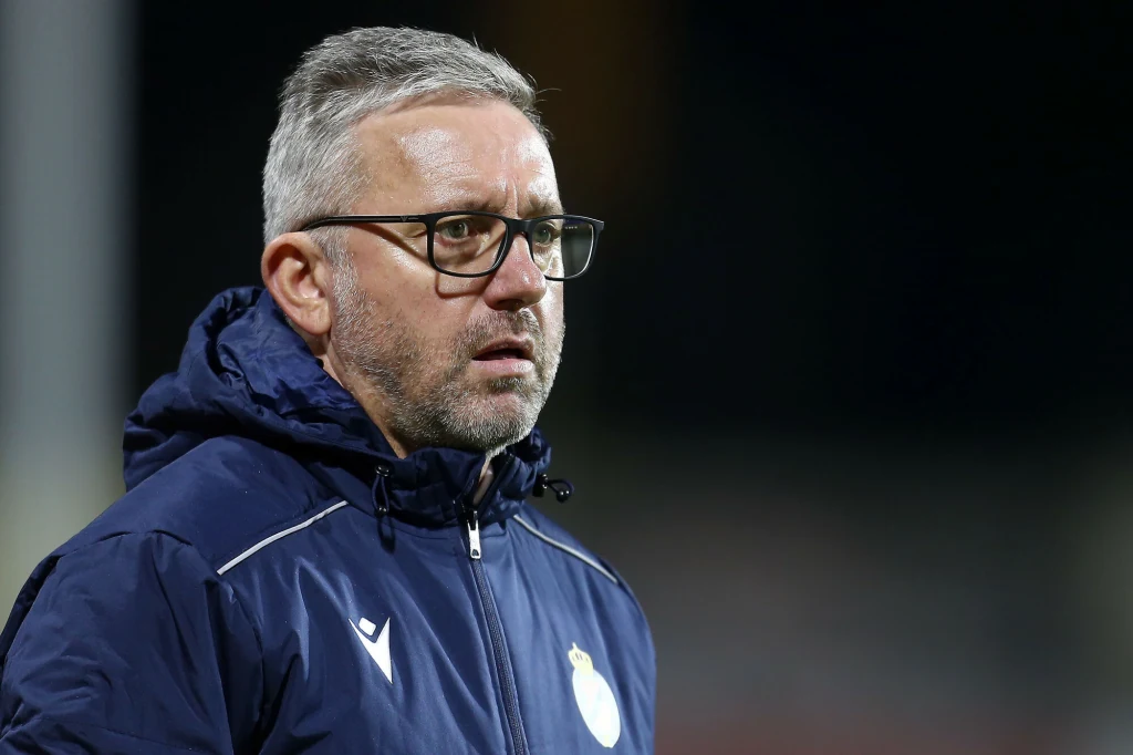 Mężczyzna w średnim wieku z siwymi włosami i brodą, noszący okulary oraz sportową kurtkę z emblematem klubu, stoi na zewnątrz w nocy i patrzy w bok z poważnym wyrazem twarzy.